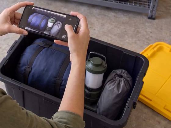 Hands holding a phone over an open bin to photograph what’s inside: a sleeping bag, a camping lantern and a stuff sack