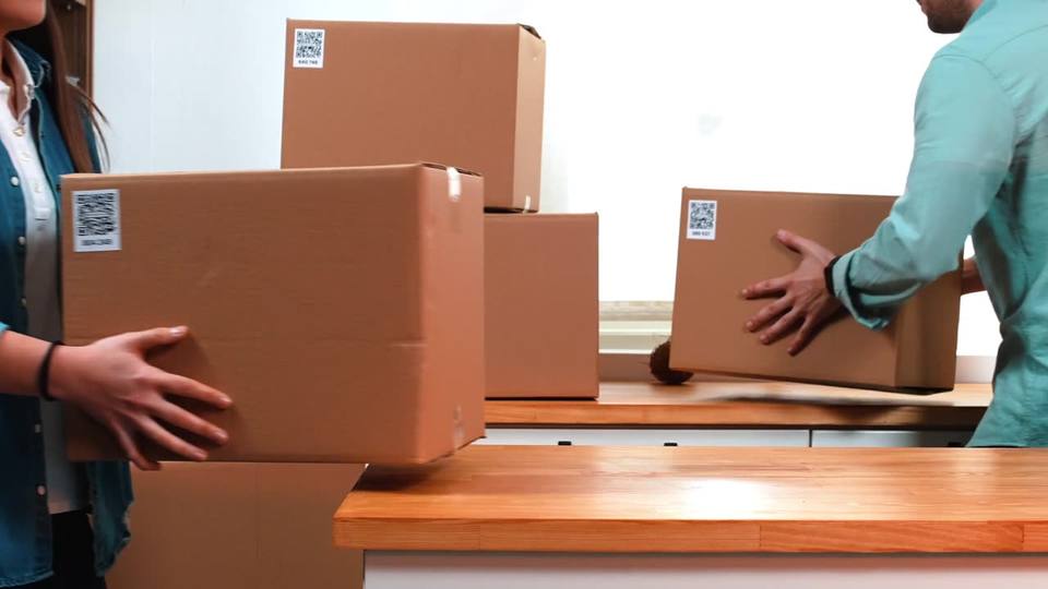 Two people carry cardboard boxes through a kitchen, each box with a QR label stuck on its side