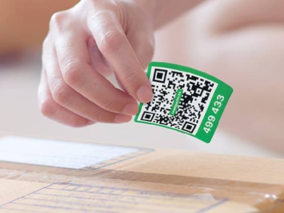 A hand holding a green QR code label, with its number printed under the code, just above a cardboard box