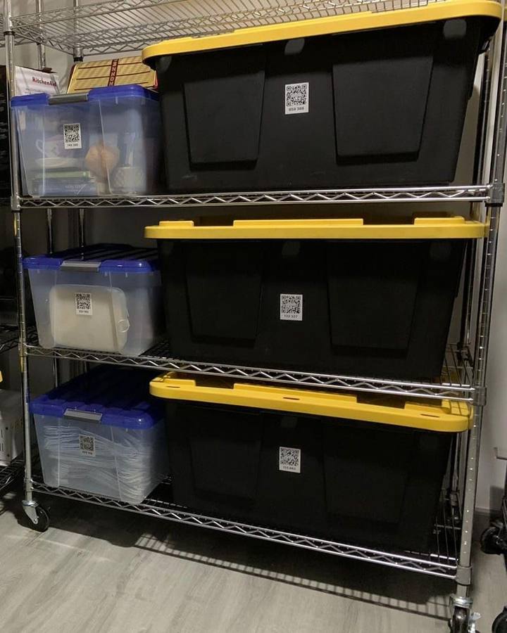 Black and clear plastic storage bins with yellow and blue lids on a metal shelving rack, each with a small QR label on its front