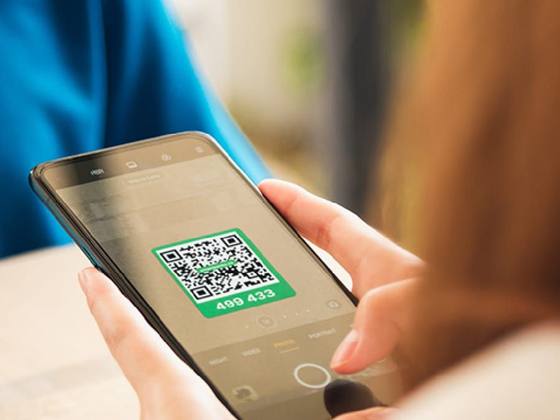 A hand holding a phone with a green QR label on the camera screen, with the same label on the box below